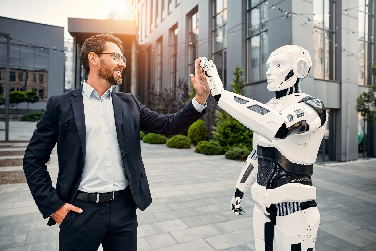 A male businessman in a business suit standing with a robot against the background of a building giving high fives to each other. The future with artificial intelligence.